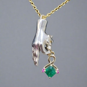 May include: A silver pendant necklace with a hand holding a gold charm. The charm features a square, emerald-green gemstone with pink accents, suspended from a gold chain. The necklace is set against a grey background.