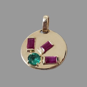 May include: A gold pendant with a circular design, featuring three rectangular ruby gemstones and one round emerald gemstone. The pendant has a loop at the top for a chain.