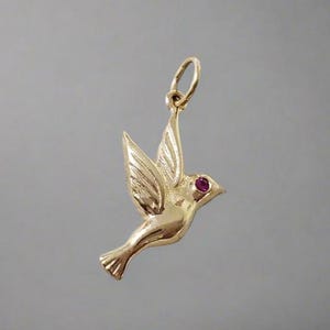 May include: A gold-coloured bird pendant with a ruby-coloured eye. The bird is depicted in flight with its wings spread. The pendant has a loop at the top for attaching to a chain. Jewellery.