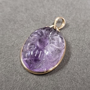 Op de afbeelding: Een hanger van paarse amethyst, gesneden met een bloemmotief, ingelijst in een gouden metalen frame.