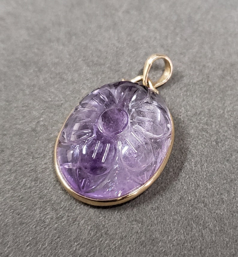 Op de afbeelding: Een hanger van paarse amethyst, gesneden met een bloemmotief, ingelijst in een gouden metalen frame.