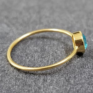 May include: A gold ring with a round, light blue gemstone. The ring band is thin and smooth, and the gemstone is set in a square bezel. The ring is shown on a gray surface.
