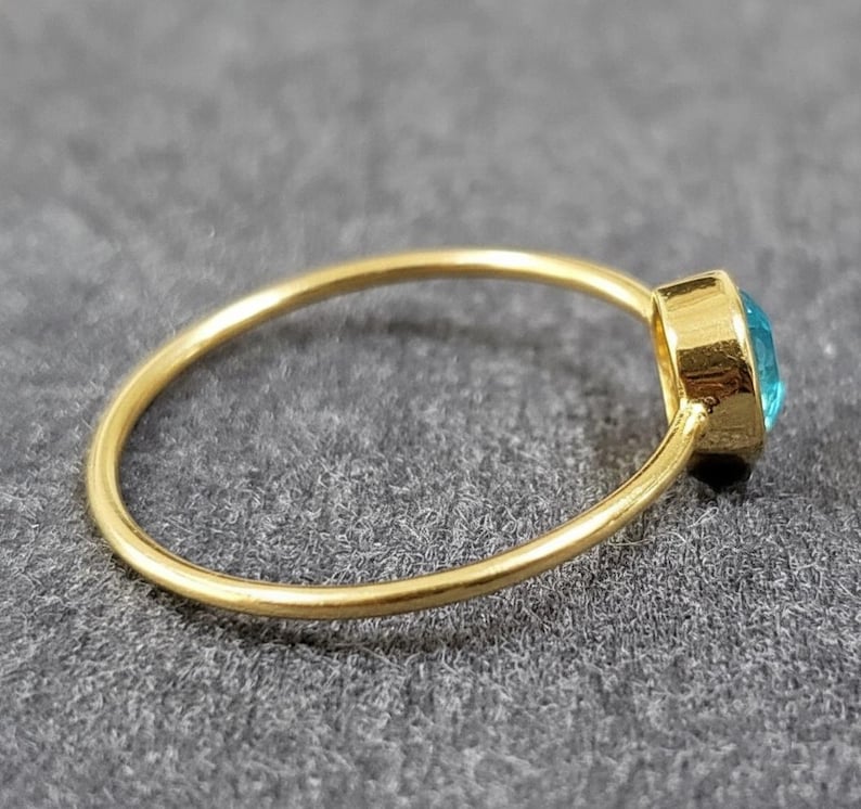 May include: A gold ring with a round, light blue gemstone. The ring band is thin and smooth, and the gemstone is set in a square bezel. The ring is shown on a gray surface.
