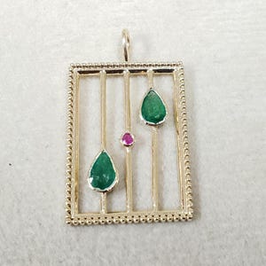 May include: A gold-colored pendant with a rectangular frame and vertical bars. Two teardrop-shaped green gemstones and one small round pink gemstone are set within the frame. The frame has a beaded border and a loop for a chain.