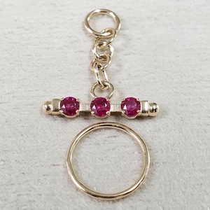May include: A gold-toned necklace clasp with three red gemstones set in a horizontal bar, connected by a chain to a circular ring. The clasp is displayed on a light gray surface.