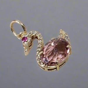 Può includere: Un ciondolo a forma di cigno in oro con una pietra preziosa rosa e dettagli trasparenti. Il ciondolo ha un anello per attaccarlo a una catena.