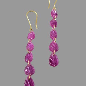 May include: A pair of dangle earrings featuring carved magenta leaf-shaped gemstones suspended from gold-colored hooks. Each earring has five leaves connected by small gold links, creating a delicate, flowing design.