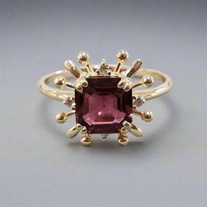 May include: A gold ring with a square, pink gemstone surrounded by smaller diamonds and a burst of gold accents.