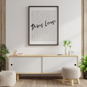 May include: A black and white framed print with the lyrics to the song "Drivers License" by Olivia Rodrigo. The lyrics are printed in a bold, sans-serif font. The print is hanging on a wall above a wooden cabinet.