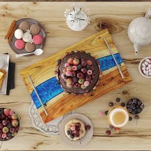 May include: A wooden serving tray with a blue epoxy resin inlay, featuring a chocolate cake topped with fresh berries and cherries. The tray is surrounded by other food items, including macarons, toast, and a cup of hot chocolate.