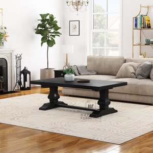 May include: Black wooden coffee table with a rectangular top and turned legs. The table is sitting on a cream-colored rug with a wavy pattern. The table is in a living room setting with a beige sofa and a large green plant.