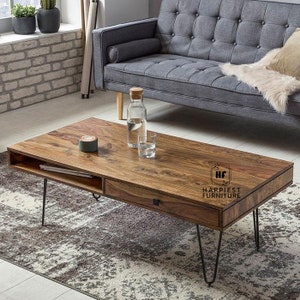 Könnte beinhalten: Ein rechteckiger Holz-Couchtisch mit einer Schublade und einem Regal. Der Tisch hat schwarze Haarnadelbeine und eine dunkelbraune Holzoptik. Der Tisch steht auf einem gemusterten Teppich vor einem grauen Sofa.