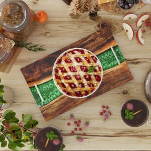 May include: A wooden serving tray with a green epoxy resin inlay. The tray features a cherry pie with a lattice crust and a red filling. The tray is surrounded by small red berries and green leaves.