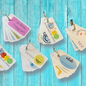 May include: Set of five small card decks with key rings. The decks are white with colourful designs and text. The decks include "My Anxiety Busting Cards", "My Behaviour Cards", "My Grounding Cards", "Excited", and "Inhale for a count of 5".