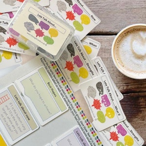 May include: A set of colourful conversation cards with the text "Lets Talk About..." and different prompts for conversation starters. The cards are arranged on a wooden table with a cup of coffee.