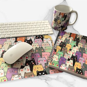 May include: A white keyboard, mouse, mug, mousepad, and spiral notebook are arranged on a marble surface. The mug, mousepad, and notebook feature a repeating pattern of colourful cartoon cats. The mouse is white.