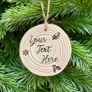 May include: A round wooden Christmas ornament with the text "Your Text Here" engraved in a decorative font. The ornament features a snowflake, bow, and holly design. It hangs from a natural twine string against a backdrop of green pine needles.