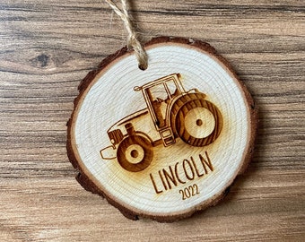 Może przedstawiać: Drewniana ozdoba choinkowa w kształcie plastra drewna. Ozdoba przedstawia grawerowany laserowo wzór traktora z imieniem "LINCOLN" i rokiem "2022". Do zawieszenia przymocowana jest pętla ze sznurka. Średnica ok. 8 cm.