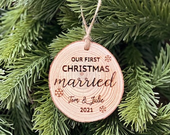 May include: A wooden Christmas ornament with the text "OUR FIRST CHRISTMAS married" and the names "Tom & Julie" with the year "2021" engraved on it. The ornament is round, made of light-colored wood, and has a twine loop for hanging. Snowflakes are also engraved.