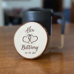 Puede incluir: Un posavasos de madera con borde natural, grabado con los nombres "Alex" y "Brittany", dos corazones entrelazados y la fecha "10-10-20". Una taza negra y beige est&aacute; en el fondo. El posavasos est&aacute; sobre una superficie de madera.