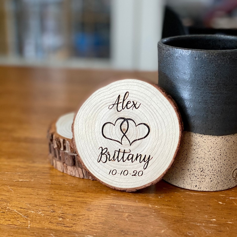 Puede incluir: Posavasos de madera redondo con borde de corteza, grabado con los nombres "Alex" y "Brittany", dos corazones entrelazados y la fecha "10-10-20". Una taza de cer&aacute;mica negra est&aacute; detr&aacute;s del posavasos sobre una superficie de madera.