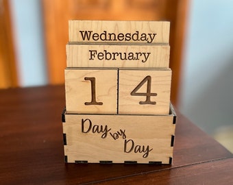Könnte beinhalten: Holzblockkalender mit dem Datum 14. Februar, Mittwoch. Die Basis des Kalenders ist mit den Worten "Tag für Tag" graviert.