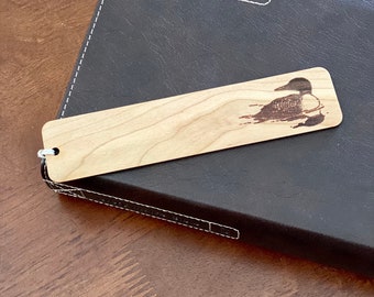 Könnte beinhalten: Ein Holz-Lesezeichen mit einem lasergravierten Bild eines Seetaucher auf einem See. Das Lesezeichen ist hellbraun und hat eine natürliche Holzmaserung. Der Seetaucher ist schwarz-weiß mit einem roten Auge. Das Lesezeichen liegt auf einem braunen ledergebundenen Notizbuch.