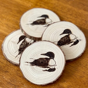 May include: Set of four wooden coasters featuring a black and white image of a loon bird. The coasters are round and have a natural wood finish.