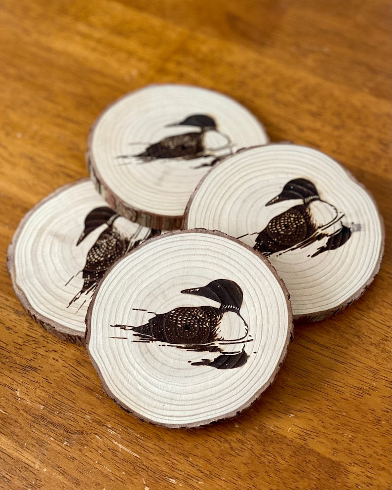 May include: Set of four wooden coasters featuring a black and white image of a loon bird. The coasters are round and have a natural wood finish.
