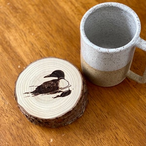 May include: A wooden coaster with a black and white image of a loon, a type of bird, on it. The coaster is sitting on a wooden table next to a white and brown speckled mug.