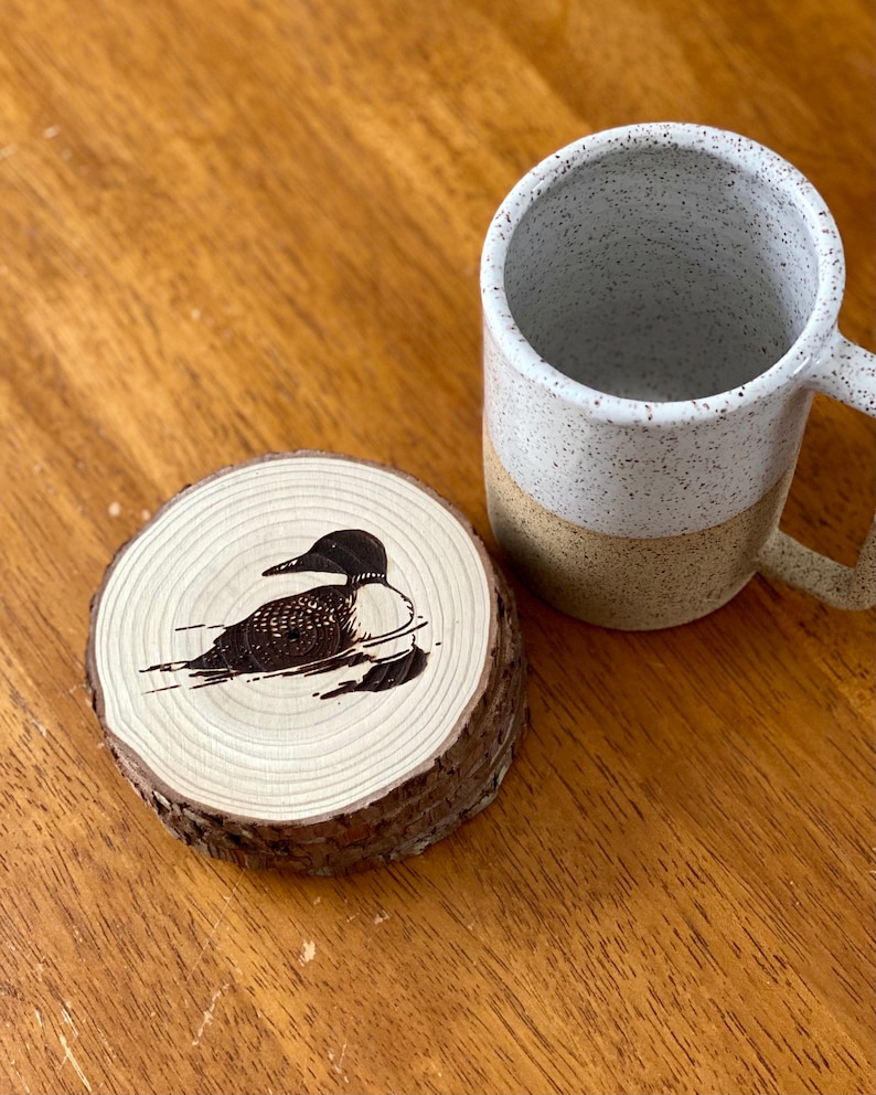 May include: A wooden coaster with a black and white image of a loon, a type of bird, on it. The coaster is sitting on a wooden table next to a white and brown speckled mug.