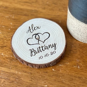 Puede incluir: Un posavasos de madera con borde de corteza natural. El posavasos presenta los nombres "Alex" y "Brittany" con dos corazones entrelazados y la fecha "10-10-20" grabada en escritura negra. Una taza de cer&aacute;mica en blanco y negro est&aacute; en el fondo.