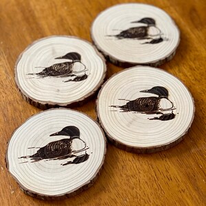 May include: Four wooden coasters with a black and white image of a loon bird on each. The coasters are made of natural wood and have a rustic look.