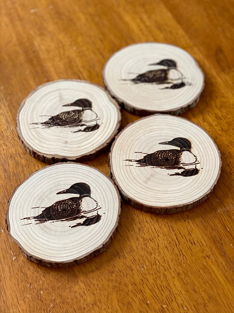 May include: Four wooden coasters with a black and white image of a loon bird on each. The coasters are made of natural wood and have a rustic look.