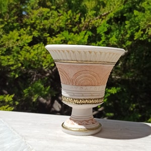 May include: A white and pink ceramic planter with gold accents and a decorative design. The planter has a wide, round top and a narrow base.