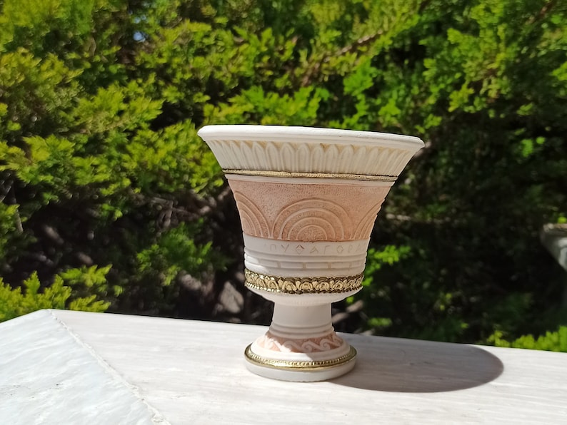 May include: A white and pink ceramic planter with gold accents and a decorative design. The planter has a wide, round top and a narrow base.