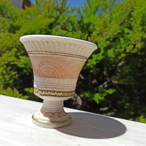 May include: A small, white ceramic planter with gold accents. The planter has a wide, round top and a narrow base. It features a decorative design of arches and a gold band around the top.