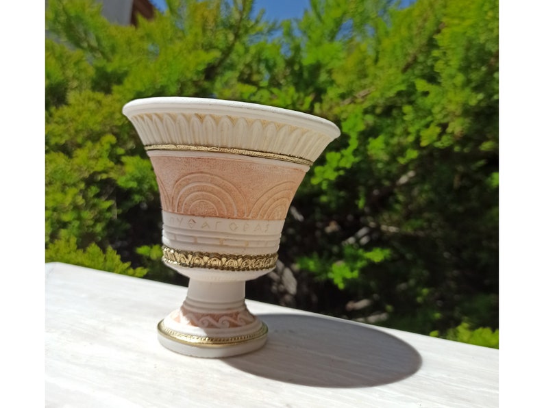 May include: A small, white ceramic planter with gold accents. The planter has a wide, round top and a narrow base. It features a decorative design of arches and a gold band around the top.