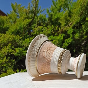 May include: A white and pink ceramic vase with gold accents and a decorative design. The vase is lying on its side on a white surface.