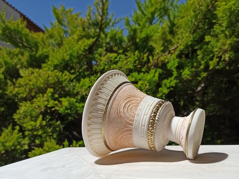 May include: A white and pink ceramic vase with gold accents and a decorative design. The vase is lying on its side on a white surface.