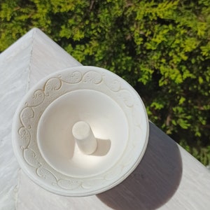 May include: White ceramic ring holder with a decorative design around the edge. The holder has a small, round, white pedestal in the center.