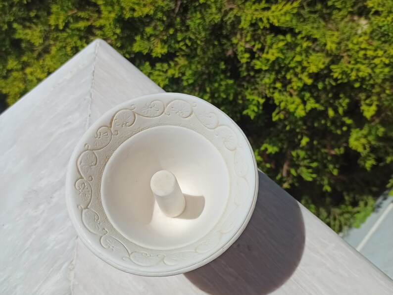 May include: White ceramic ring holder with a decorative design around the edge. The holder has a small, round, white pedestal in the center.