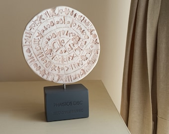 Puede incluir: Una réplica de cerámica blanca del Disco de Festos, un artefacto misterioso de la civilización minoica, sobre un soporte negro con la inscripción "Disco de Festos, Segundo Milenio a. C."