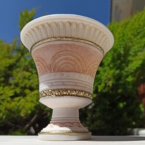 May include: A white ceramic planter with gold accents and a pink and white design. The planter has a wide, round top and a narrow base. The planter is decorated with a Greek key pattern and the word "AGAPE" in Greek letters.