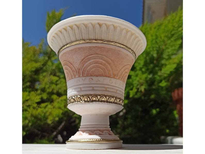 May include: A white ceramic planter with gold accents and a pink and white design. The planter has a wide, round top and a narrow base. The planter is decorated with a Greek key pattern and the word "AGAPE" in Greek letters.