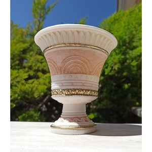 May include: A white and pink ceramic planter with gold accents. The planter has a decorative design with arches and a gold band around the top.
