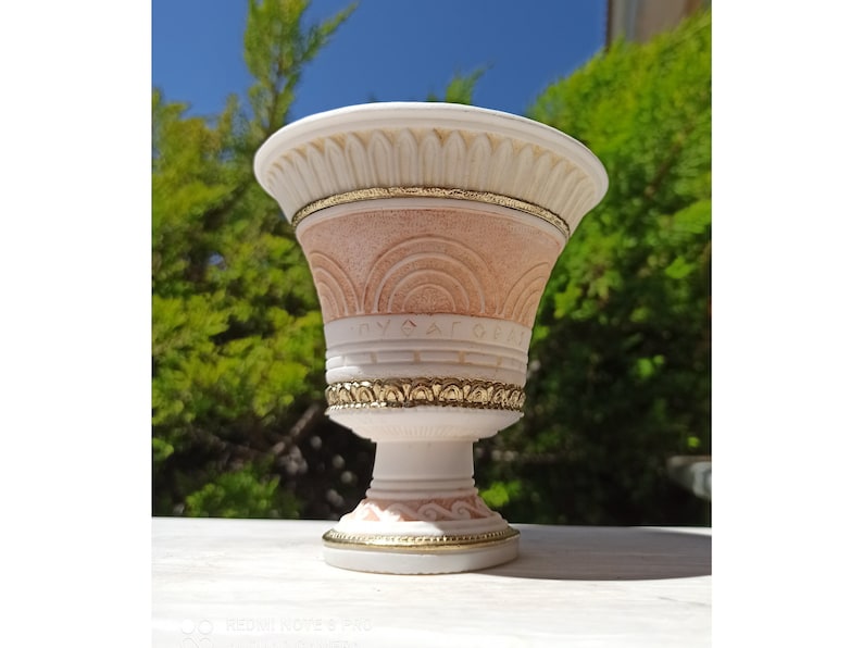 May include: A white and pink ceramic planter with gold accents. The planter has a decorative design with arches and a gold band around the top.
