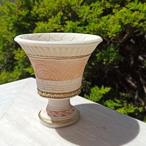 May include: A white ceramic vase with gold accents and intricate designs. The vase has a wide, round base and a narrow neck. It is decorated with a pattern of circles and lines.