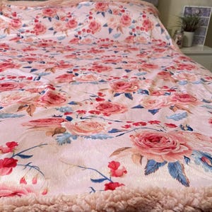 Könnte beinhalten: Eine rosa Fleece-Decke mit einem Blumenmuster aus rosa Rosen und blauen Blättern. Die Decke hat eine flauschige, weiche Textur.