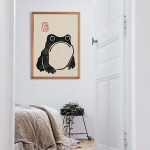 Large framed Japanese frog illustration hangs in a bright minimalist bedroom with natural linens and soft wooden flooring. The quirky frog and red seal detail bring a touch of humor and refined calm, blending seamlessly with Japandi and zen decor.